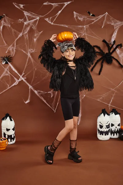 Menina pré-adolescente feliz segura abóbora na cabeça com as mãos, fundo marrom com teia de aranha, Halloween — Fotografia de Stock