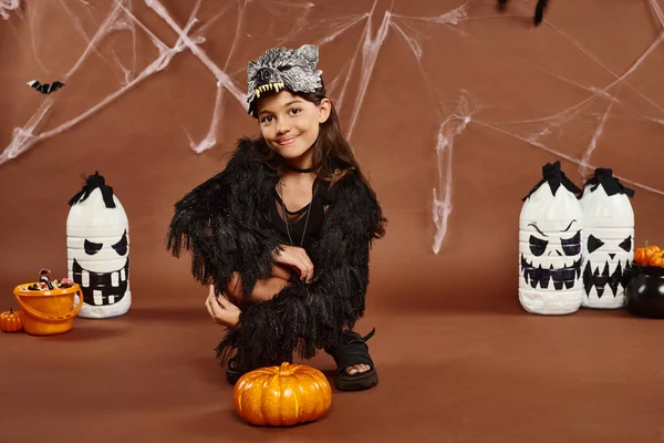 Cheerful preteen girl squatting near pumpkin with lanterns and spiderweb on backdrop, Halloween — Stock Photo