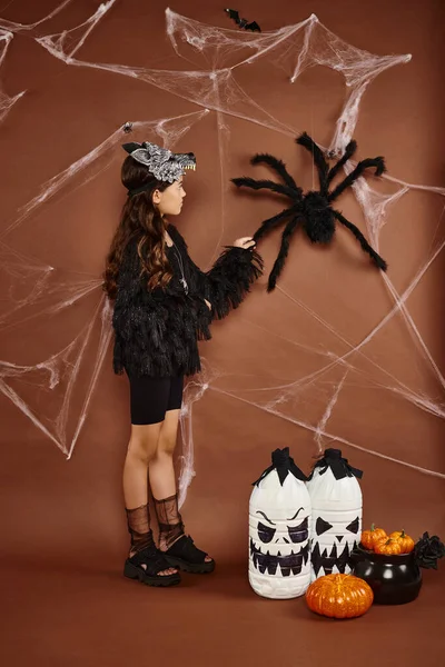 Мила дівчина в вовчій масці і чорний одяг торкається павука на коричневому тлі, концепція Хеллоуїна — стокове фото