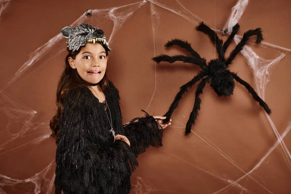 Nahaufnahme verängstigtes Mädchen in Wolfsmaske und schwarzem Gewand, das Spinne auf braunem Hintergrund berührt, Halloween — Stockfoto