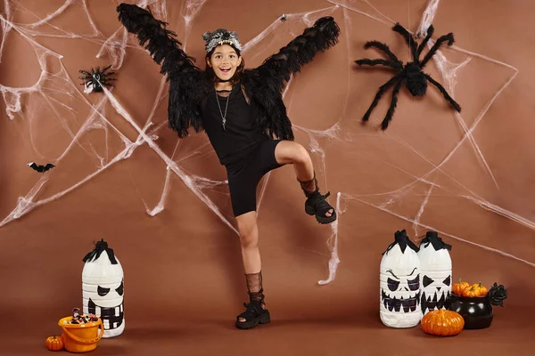 Chica feliz con los brazos abiertos en traje negro de pie en una pierna con telaraña en el telón de fondo, Halloween - foto de stock