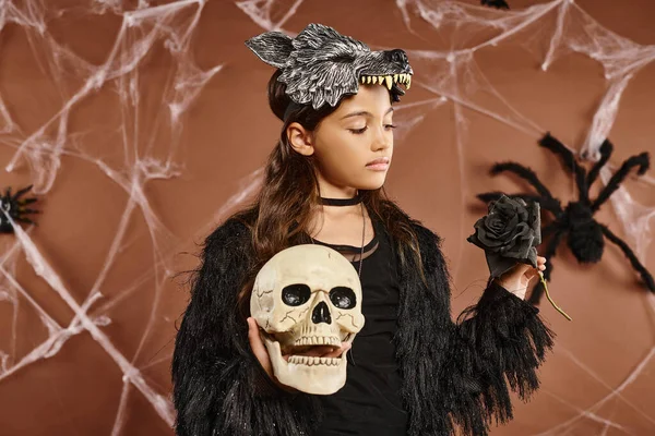 Cerca de niña preadolescente sostiene cráneo y rosa negra en sus manos usando máscara de lobo, concepto de Halloween - foto de stock