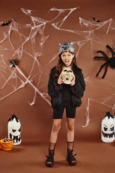 Preteen girl grimacing and holding skull on brown backdrop with spiderweb and lanterns, Halloween — Stock Photo
