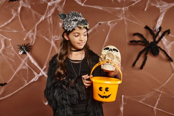 Sorrindo menina pré-adolescente segurando crânio e balde de doces, fundo marrom com teia de aranha, Halloween — Fotografia de Stock