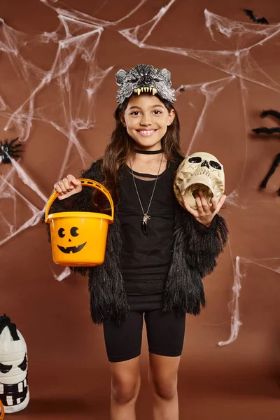 Nahaufnahme lächelndes Mädchen mit Totenkopf und Eimer voller Süßigkeiten, brauner Hintergrund mit Netz, Halloween — Stockfoto