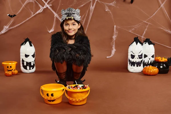 Preteen kid squatting down near buckets of sweets with lanterns and pumpkins on backdrop, Halloween — Stock Photo