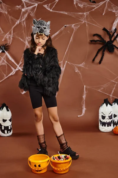 Reflexiva niña preadolescente en traje de piel sintética negro posando cerca de cubos de dulces, Halloween - foto de stock