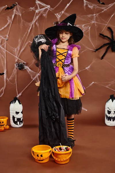 Fille souriante en chapeau de sorcière avec toile d'araignée maquillage tenant jouet effrayant près des seaux de citrouille, Halloween — Photo de stock