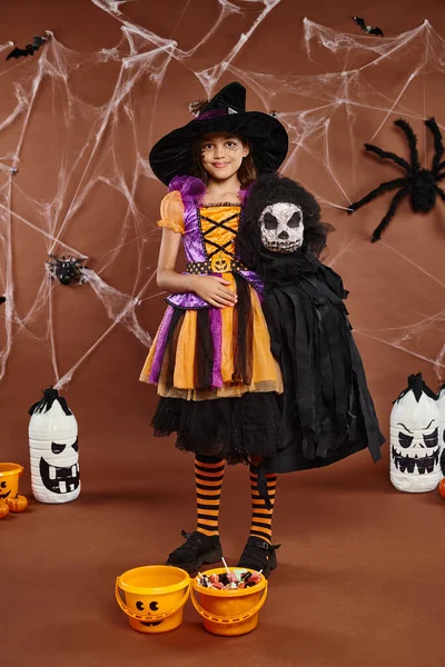Menina alegre em chapéu de bruxa com maquiagem cobweb segurando brinquedo assustador no fundo marrom, Halloween — Fotografia de Stock