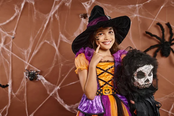 Close up preteen girl in witch hat with spooky toy touching her cheek and smiling, Halloween — Stock Photo