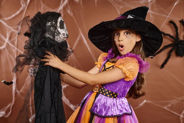 Primer plano chica sorprendida en sombrero de bruja y araña maquillaje sostiene juguete de miedo, fondo marrón, Halloween - foto de stock