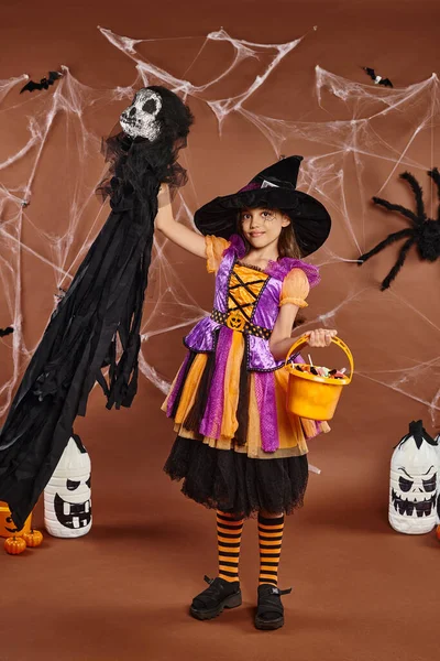 Happy kid in witch hat with spiderweb makeup holding bucket of sweets and spooky toy, Halloween — Stock Photo