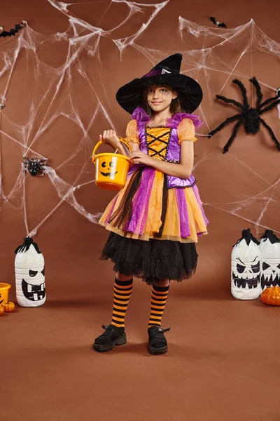 Niedlichen Preteen in Hexenhut hält Eimer mit Süßigkeiten auf braunem Hintergrund mit Spinnennetzen, Halloween — Stockfoto