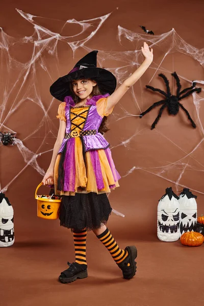 Criança pré-adolescente sorridente segura balde de doces e acenando, fundo marrom com teia de aranha, Halloween — Fotografia de Stock