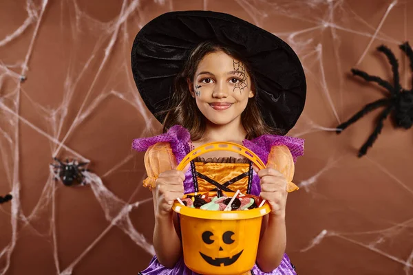 Happy girl in witch hat with spiderweb makeup holding bucket of sweets on brown with spider net — Stock Photo
