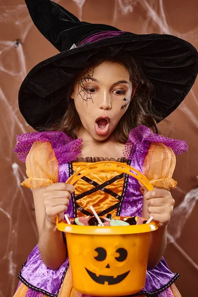 Chaval en sombrero de bruja y disfraz de Halloween mirando dulces en cubo sobre fondo marrón - foto de stock