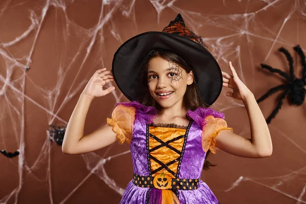 Menina alegre no traje de Halloween ajustando chapéu de bruxa e olhando para a câmera no fundo marrom — Fotografia de Stock