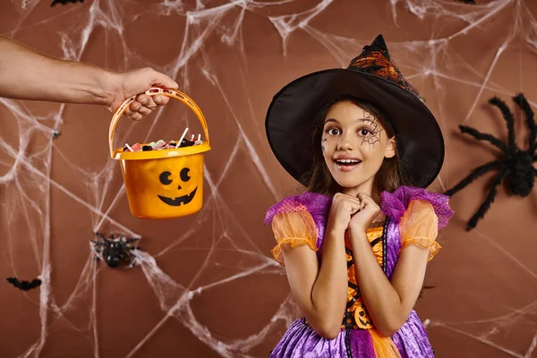 Amazed girl in witch hat and Halloween costume looking at camera near hand holding sweets in bucket — Stock Photo