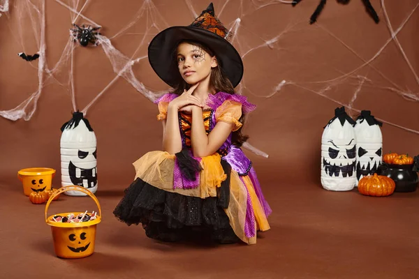 Girl in witch hat and Halloween costume looking at camera near sweets in bucket and spooky decor — Stock Photo