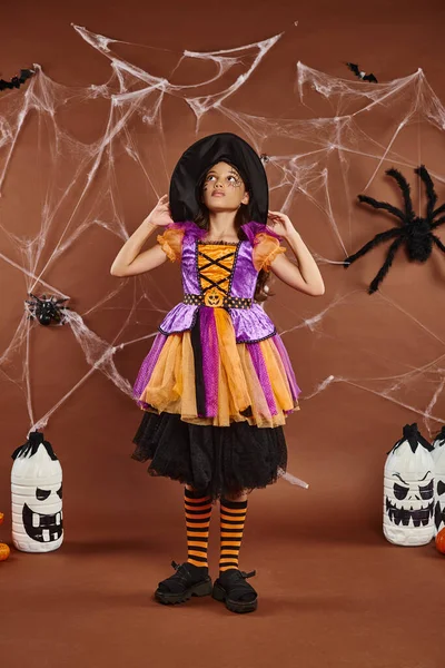 Chica en sombrero de bruja y disfraz de Halloween de pie cerca de la decoración espeluznante y telarañas en el fondo marrón - foto de stock