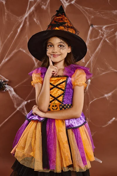 Menina feliz em chapéu de bruxa e traje de Halloween perto de teias de aranha no fundo marrom, estação assustadora — Fotografia de Stock