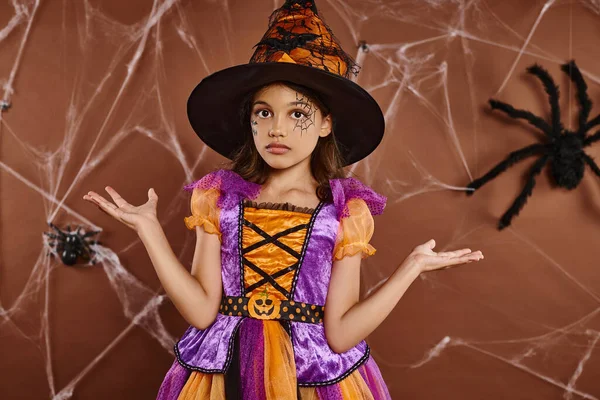Confused girl in witch hat and Halloween costume near cobwebs on brown backdrop, spooky season — Stock Photo