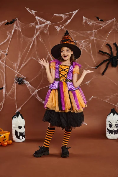 Menina feliz em chapéu de bruxa e traje de Halloween gestos perto de teias de aranha em marrom, estação assustadora — Fotografia de Stock