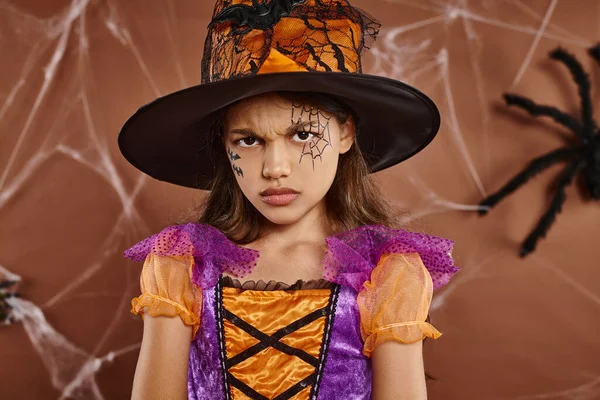 Menina descontente em chapéu de bruxa e traje de Halloween franzindo a testa em fundo marrom, estação assustadora — Fotografia de Stock