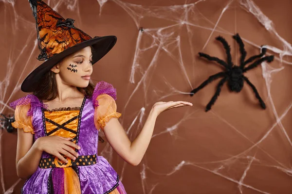 Girl in witch hat and Halloween costume pointing at fake spider on brown background, spooky season — Stock Photo