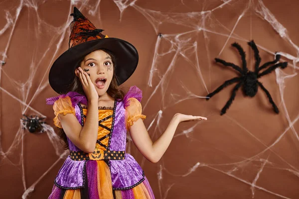 Fille étonnée en chapeau de sorcière et costume d'Halloween pointant vers fausse araignée sur fond brun, effrayant — Photo de stock