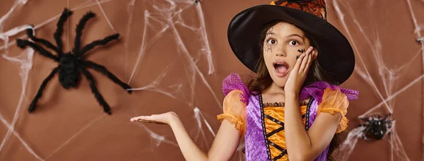 Amazed girl in witch hat and Halloween costume pointing at fake spider on brown background, banner — Stock Photo