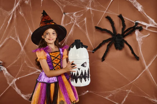 Fille heureuse en chapeau de sorcière et costume d'Halloween debout avec décor effrayant sur fond brun — Photo de stock