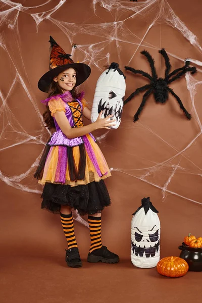 Happy girl in witch hat and Halloween costume standing with diy spooky decor on brown background — Stock Photo