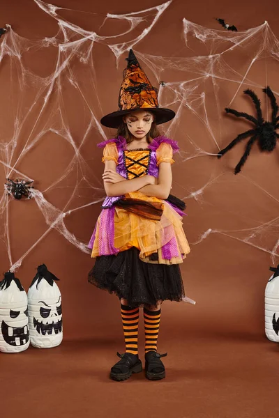 Sad girl in witch hat and Halloween costume standing with folded arms on brown backdrop, cobwebs — Stock Photo