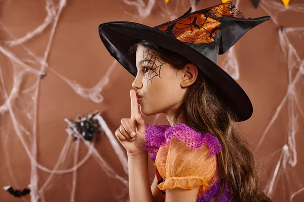 Cute little girl in Halloween witch costume and pointed hat showing shh on brown backdrop, side view — Stock Photo