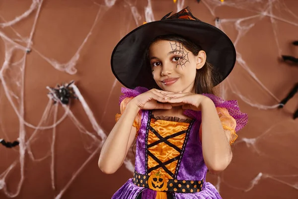 Linda chica en disfraz de bruja de Halloween y sombrero puntiagudo sonriendo sobre fondo marrón, temporada espeluznante - foto de stock