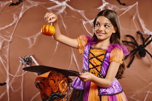 Positive child in Halloween witch costume holding pumpkin near pointed hat on brown background — Stock Photo