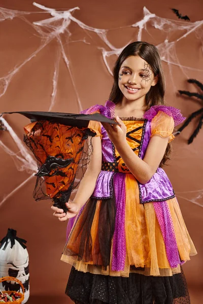 Cheerful child in Halloween witch costume holding pointed hat on brown background, spooky season — Stock Photo