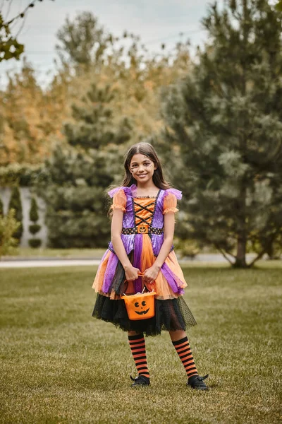 Niedliches Kind im Halloween-Kostüm, das mit einem Eimer süßer Bonbons auf grünem Gras steht, Mädchen im Kleid — Stockfoto