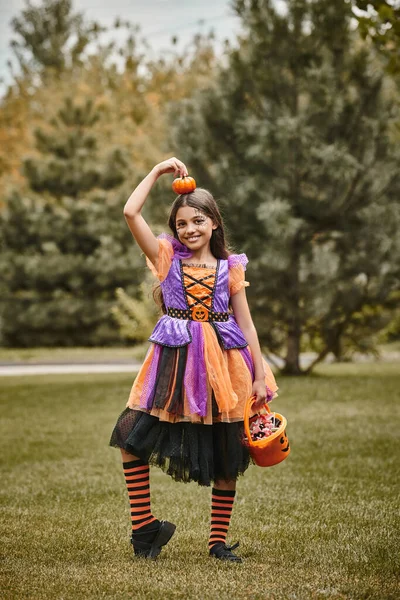 Happy girl in Halloween costume holding pumpkin and bucket of candies on green grass, kid in dress — Stock Photo