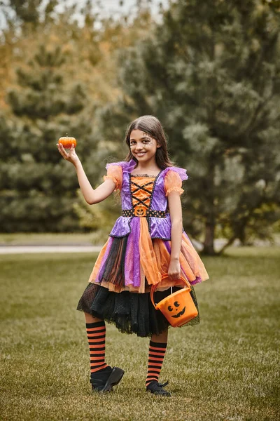 Cheerful girl in Halloween costume holding pumpkin and bucket of candies on green grass — Stock Photo