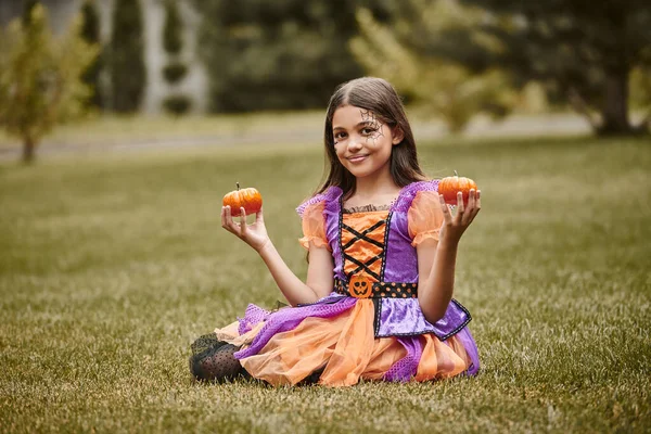 Cheerful girl in Halloween costume sitting in vibrant dress on green grass and holding tiny pumpkins — Stock Photo