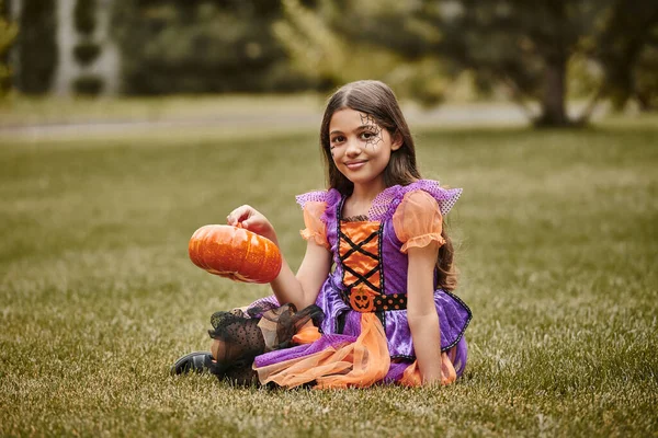 Joyful girl in Halloween costume sitting on green grass and holding decorative pumpkin — Stock Photo