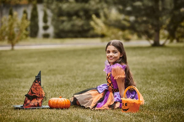 Menina feliz no traje de Halloween perto de abóbora decorativa, chapéu apontado e balde de doces na grama — Fotografia de Stock