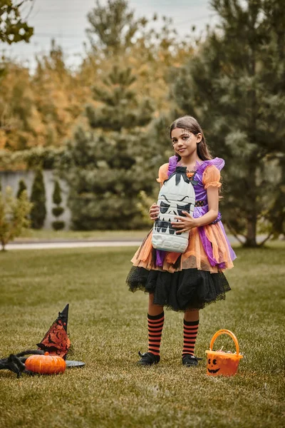 Menina no traje de Halloween segurando diy decoração assustadora perto de abóbora, chapéu apontado e balde de doces — Fotografia de Stock