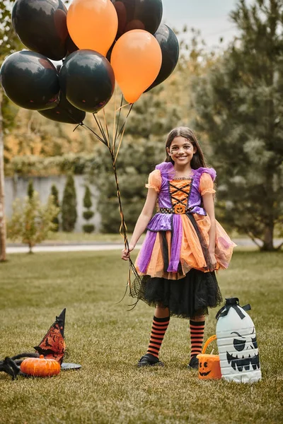 Chica feliz en disfraz de Halloween celebración de globos cerca de la calabaza, sombrero puntiagudo y balde de caramelo en la hierba - foto de stock