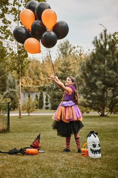 Joyful girl in Halloween costume holding balloons near pumpkin, witch hat and candy bucket on grass — Stock Photo