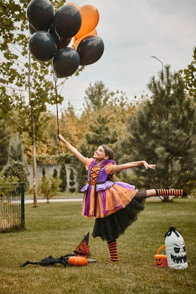 Chica alegre en traje de Halloween celebración de globos cerca de la calabaza, sombrero de bruja y cubo de dulces en la hierba - foto de stock