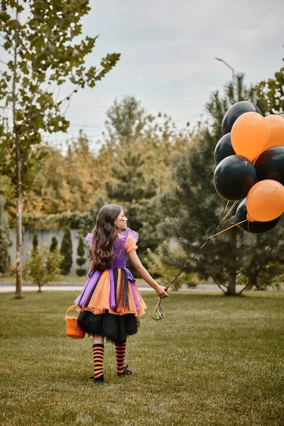 Menina bonito no traje de Halloween segurando balões e balde de doces enquanto caminhava na grama verde — Fotografia de Stock
