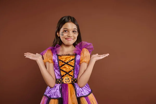 Chica confundida en disfraz de Halloween con maquillaje de tela de araña sonriendo y haciendo gestos en el fondo marrón - foto de stock